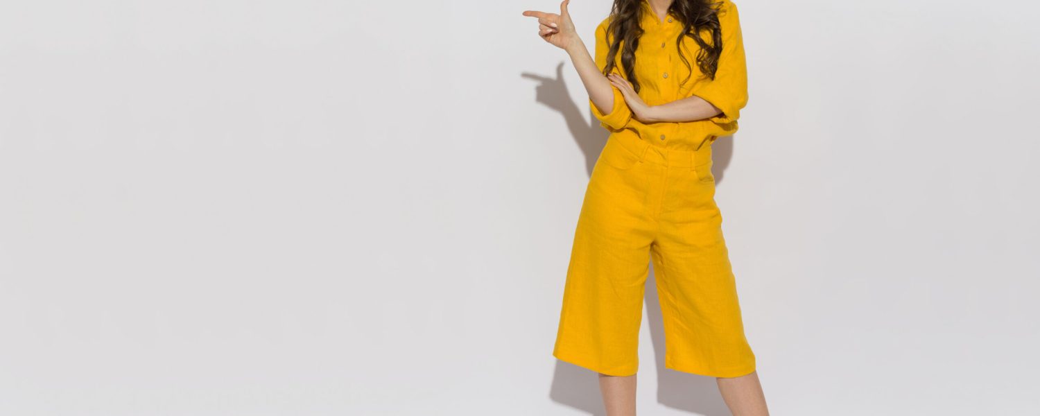 Beautiful young woman in sunglasses, yellow linen shirt and shorts standing, pointing to the side and talking. Full length studio shot.